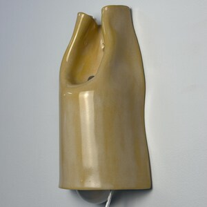 Pu&ograve; includere: Una lampada da parete in ceramica con una finitura lucida color giallo pallido. L'oggetto presenta una forma allungata, organica e asimmetrica con una sezione superiore incavata. Una lampadina &egrave; visibile in basso, collegata da un cavo trasparente.