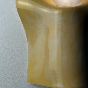 May include: A wall-mounted sconce featuring a smooth, mustard-yellow ceramic body with a curved, organic shape. A single, spherical, frosted white glass light bulb is recessed into the upper section, emitting a soft, warm glow from within.