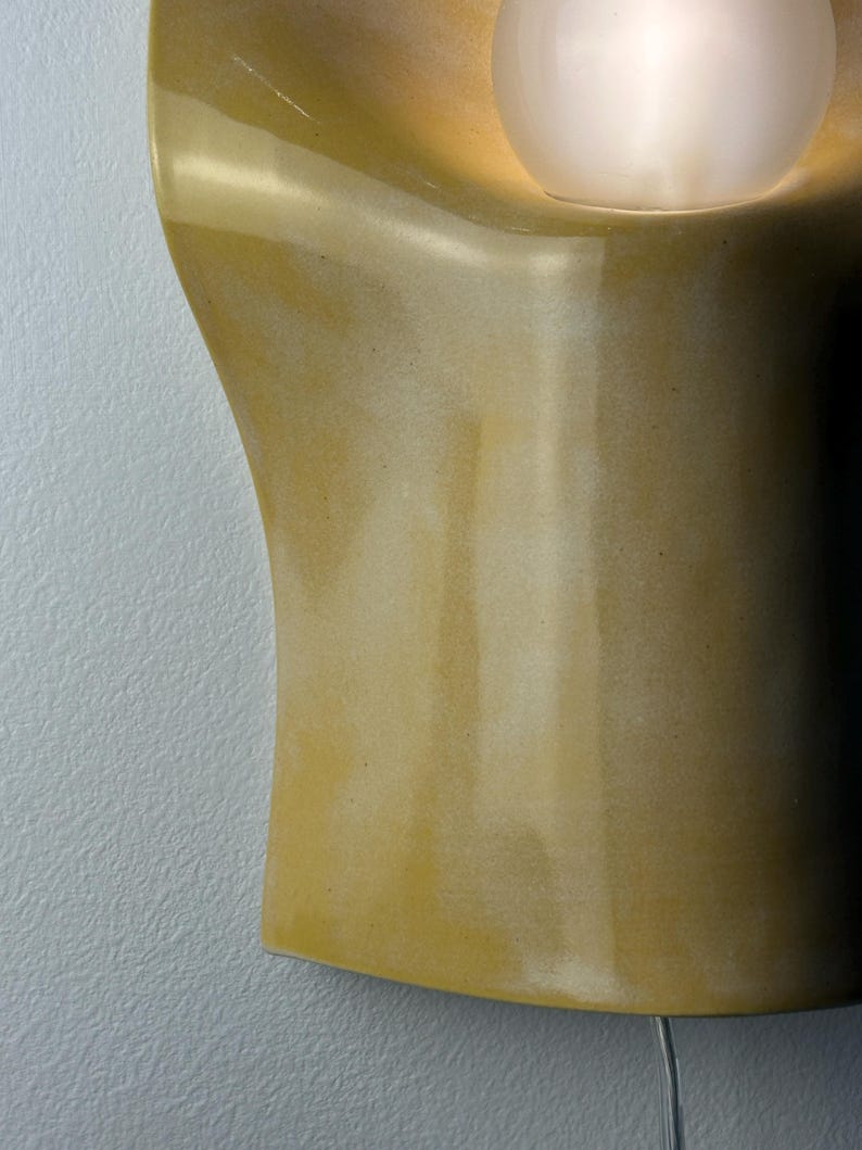 May include: A wall-mounted sconce featuring a smooth, mustard-yellow ceramic body with a curved, organic shape. A single, spherical, frosted white glass light bulb is recessed into the upper section, emitting a soft, warm glow from within.