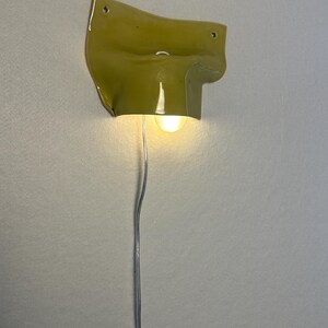 May include: A unique, wall-mounted light fixture with a textured, olive-green ceramic shade. The fixture has a bulb that emits a warm glow, and a silver wire extends downward. The design is modern and artistic, suitable for interior decor.