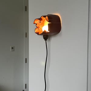 May include: A unique, brown wall sconce with an exposed light bulb. The fixture has an organic, sculpted shape and casts a warm, inviting glow. A black cord extends downward from the sconce.