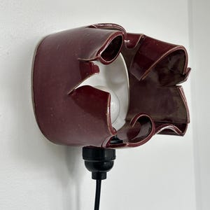 May include: A burgundy ceramic wall sconce with a unique, ruffled design. The sconce has a white interior and a black cord and bulb socket. The light fixture is mounted on a white wall.