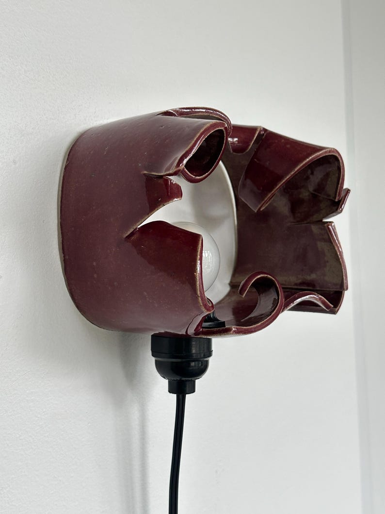 May include: A burgundy ceramic wall sconce with a unique, ruffled design. The sconce has a white interior and a black cord and bulb socket. The light fixture is mounted on a white wall.