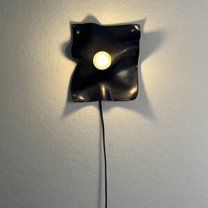 May include: A black wall-mounted lamp featuring a square, wavy, and undulating surface with a central circular light source that emits a warm glow. The fixture is connected to a thin black power cord that hangs vertically downward from the base.