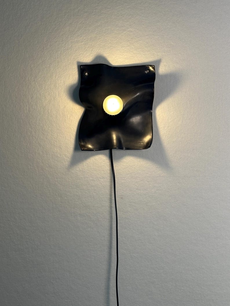 May include: A black wall-mounted lamp featuring a square, wavy, and undulating surface with a central circular light source that emits a warm glow. The fixture is connected to a thin black power cord that hangs vertically downward from the base.
