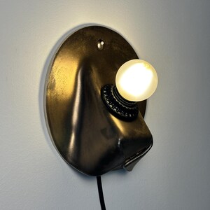 May include: A wall-mounted lamp featuring a circular, dark metallic backplate with a molded, protruding form. A single, round, glowing light bulb is secured in a black socket attached to the front, with a black power cord extending downward.