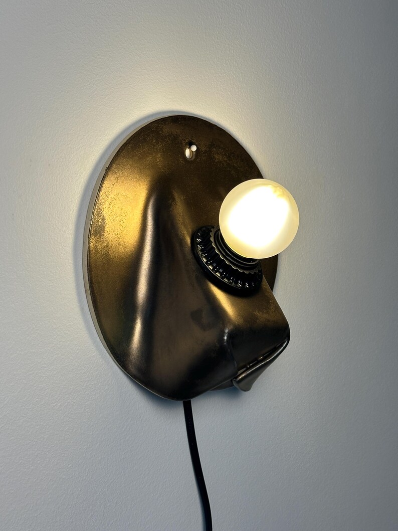 May include: A wall-mounted lamp featuring a circular, dark metallic backplate with a molded, protruding form. A single, round, glowing light bulb is secured in a black socket attached to the front, with a black power cord extending downward.