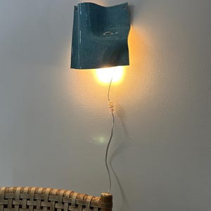May include: A wall-mounted light fixture with a glossy, speckled blue ceramic shade featuring a curved, wave-like form. The lamp emits a warm, downward-facing glow from an internal bulb, connected by a thin, clear power cord hanging vertically.