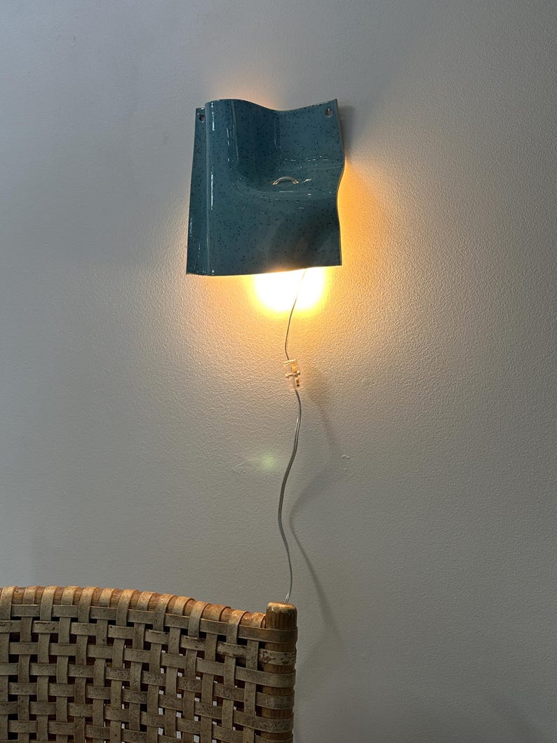 May include: A wall-mounted light fixture with a glossy, speckled blue ceramic shade featuring a curved, wave-like form. The lamp emits a warm, downward-facing glow from an internal bulb, connected by a thin, clear power cord hanging vertically.