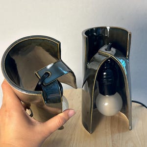 Puede incluir: Dos pantallas de l&aacute;mpara de cer&aacute;mica curvas, oscuras y brillantes con un acabado met&aacute;lico. Cada pantalla tiene una forma cil&iacute;ndrica con un lado abierto y un orificio central para un cable el&eacute;ctrico negro y un casquillo con una bombilla blanca.