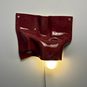 May include: A wall-mounted lamp featuring a glossy, deep red, irregularly shaped ceramic form with a fluid, draped appearance. The piece includes two small circular holes at the top corners and a silver metal ring, with a single light bulb glowing.