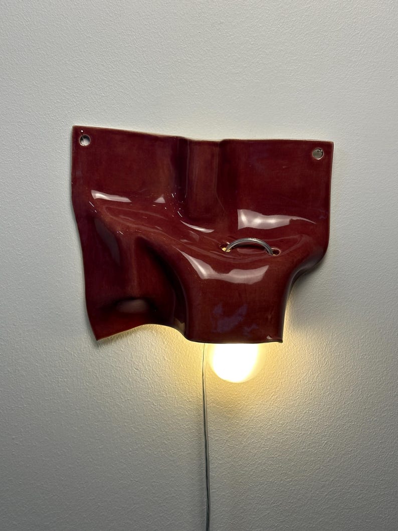 May include: A wall-mounted lamp featuring a glossy, deep red, irregularly shaped ceramic form with a fluid, draped appearance. The piece includes two small circular holes at the top corners and a silver metal ring, with a single light bulb glowing.