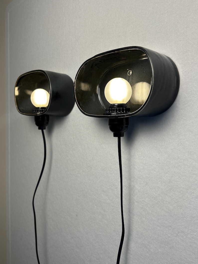 May include: Two black, oval wall sconces with exposed light bulbs. The sconces are mounted on a light grey wall, and each has a black cord. The bulbs are round and emit a warm glow.