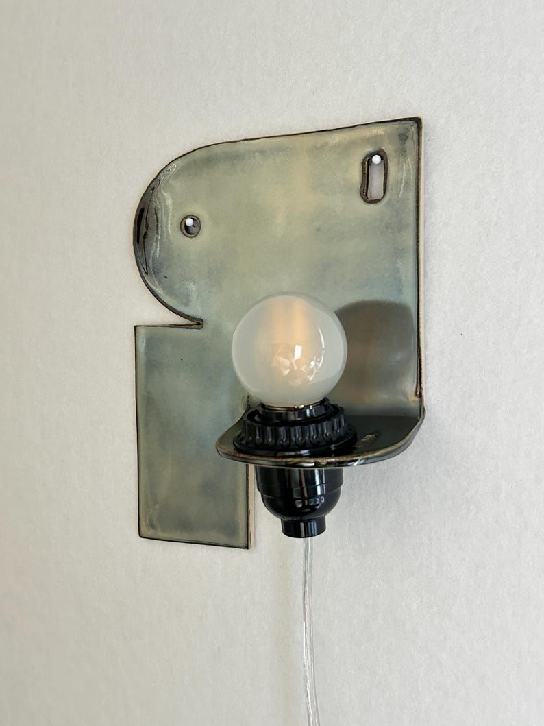 May include: A wall-mounted light fixture featuring a flat, irregularly shaped ceramic backplate with a glossy, pale green and grey glaze. A black socket holds a single round, frosted white light bulb, with a clear power cord extending downward.