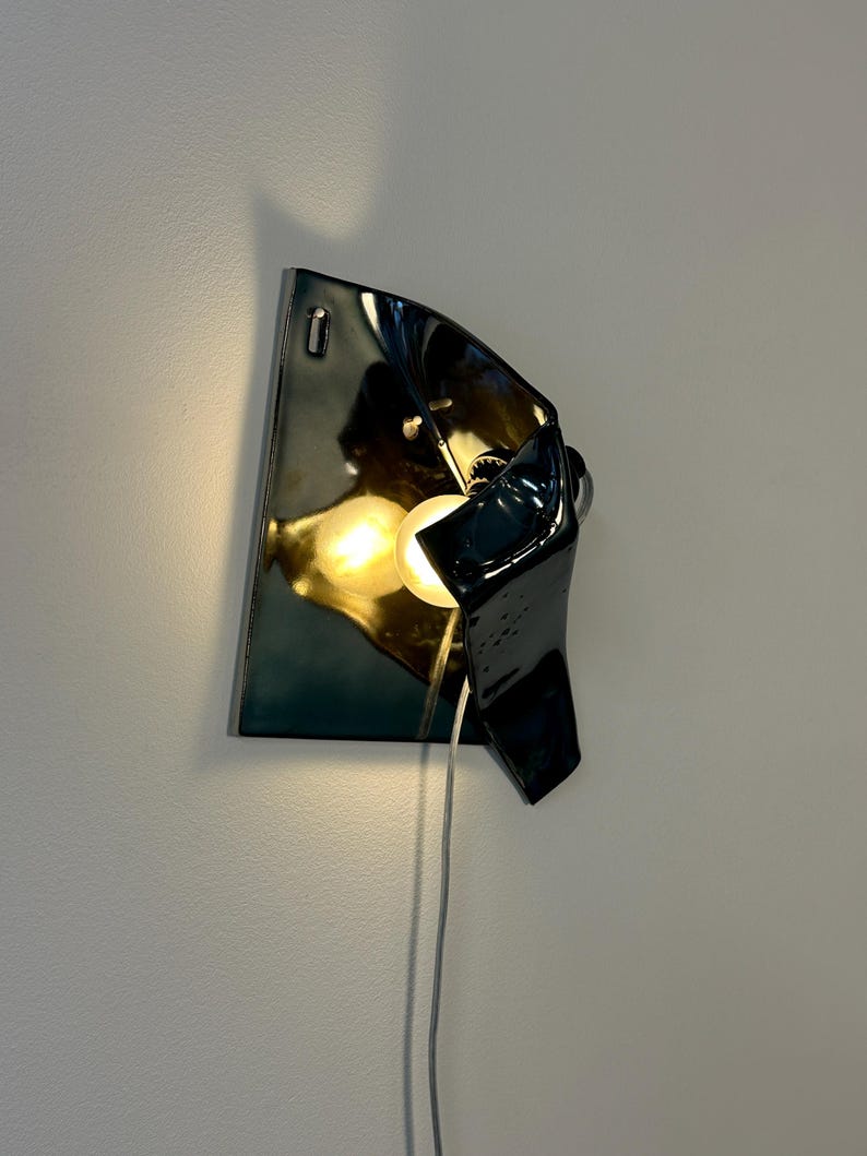 Peut inclure: Une applique murale compos&eacute;e d'une plaque en c&eacute;ramique ou en m&eacute;tal sombre et brillant, dot&eacute;e d'un design sculptural repli&eacute;. Une ampoule &agrave; lumi&egrave;re chaude est plac&eacute;e dans la courbe, diffusant une lueur douce avec un fin cordon blanc.