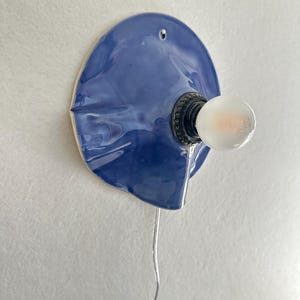 May include: A blue ceramic wall sconce with a bulb. The sconce has a unique, organic shape and a glossy finish. The bulb is round and emits a soft glow. A silver wire extends from the sconce.