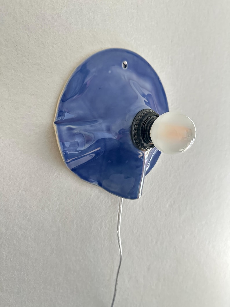 May include: A blue ceramic wall sconce with a bulb. The sconce has a unique, organic shape and a glossy finish. The bulb is round and emits a soft glow. A silver wire extends from the sconce.