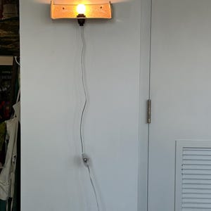 May include: A wall-mounted light fixture with a rectangular, tan-colored shade. The light bulb is illuminated, casting a warm glow. The fixture is connected to a power outlet via a thin, silver cord. The light is mounted on a white wall.