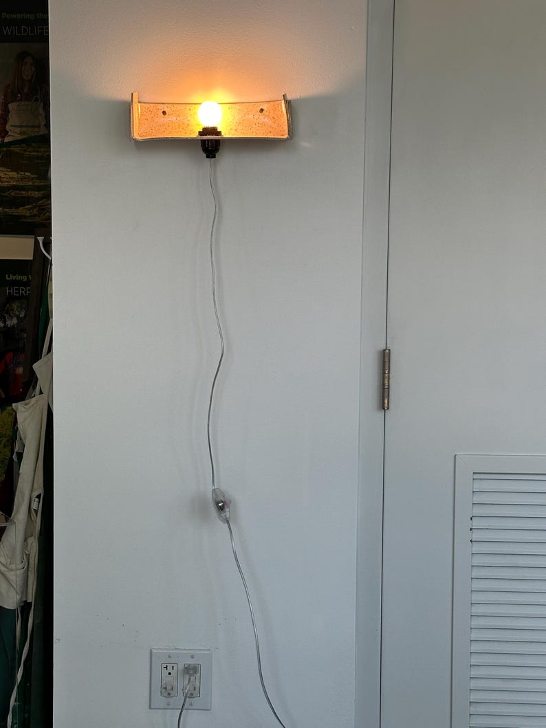 May include: A wall-mounted light fixture with a rectangular, tan-colored shade. The light bulb is illuminated, casting a warm glow. The fixture is connected to a power outlet via a thin, silver cord. The light is mounted on a white wall.