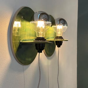 May include: Two wall-mounted light fixtures featuring circular, glossy green ceramic backplates with small horizontal shelves. Each fixture holds a clear glass globe light bulb with a silver-dipped top, black socket, and a thin, transparent power cord.