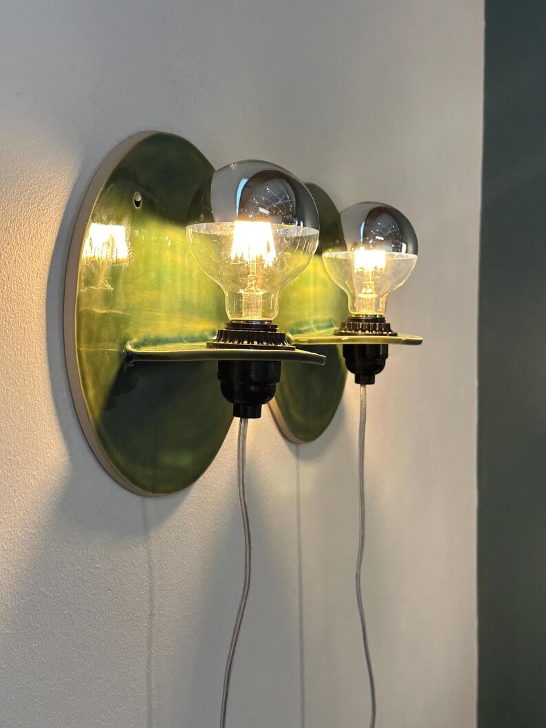 May include: Two wall-mounted light fixtures featuring circular, glossy green ceramic backplates with small horizontal shelves. Each fixture holds a clear glass globe light bulb with a silver-dipped top, black socket, and a thin, transparent power cord.