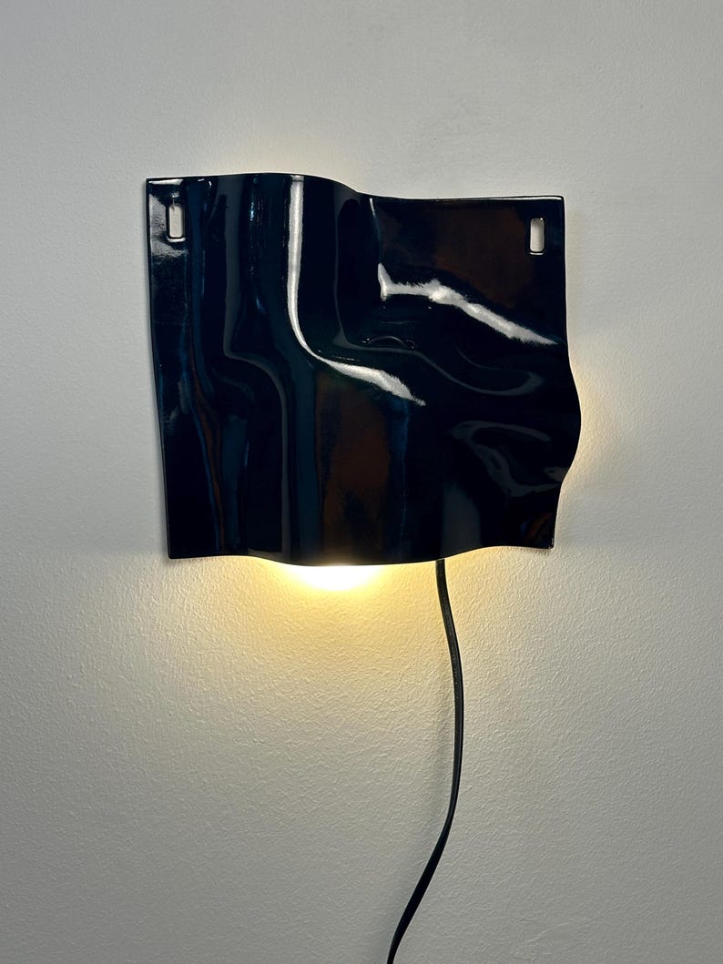 Puede incluir: Un aplique de pared de cer&aacute;mica en color azul marino oscuro brillante con una superficie ondulada. El accesorio rectangular cuenta con dos ranuras de montaje verticales en las esquinas superiores y emite una luz c&aacute;lida hacia abajo con un cable negro.