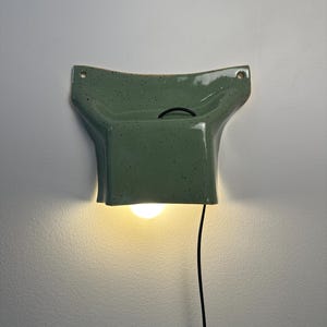 May include: A wall-mounted ceramic light fixture in a sage green speckled glaze. The piece features a flared, wide top with two circular mounting holes and a rectangular base that houses a single glowing light bulb, connected by a black cord.