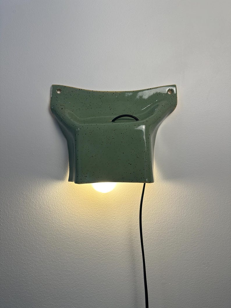May include: A wall-mounted ceramic light fixture in a sage green speckled glaze. The piece features a flared, wide top with two circular mounting holes and a rectangular base that houses a single glowing light bulb, connected by a black cord.