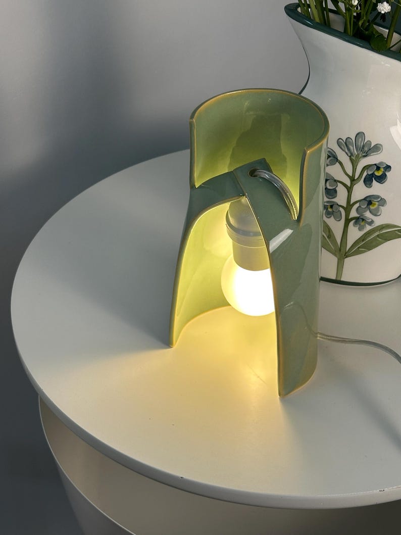 May include: A sage green ceramic table lamp with a curved, open-sided cylindrical structure. The lamp features a central white light bulb socket and a glowing bulb, with a thin white power cord extending from the base of the ceramic body.