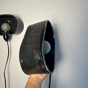 May include: Two black ceramic wall sconces with a glossy finish. Each sconce has a unique, organic shape with a bulb inside. The sconces are connected to a black electrical cord.