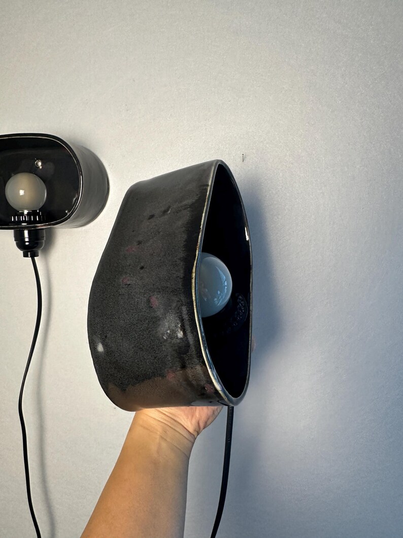 May include: Two black ceramic wall sconces with a glossy finish. Each sconce has a unique, organic shape with a bulb inside. The sconces are connected to a black electrical cord.