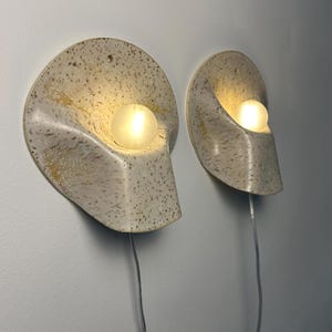 May include: Two wall-mounted sconces featuring a speckled cream and tan ceramic shell-like shade with a curved, organic form. Each fixture holds a single glowing spherical light bulb and includes a thin, clear electrical cord extending downwards.