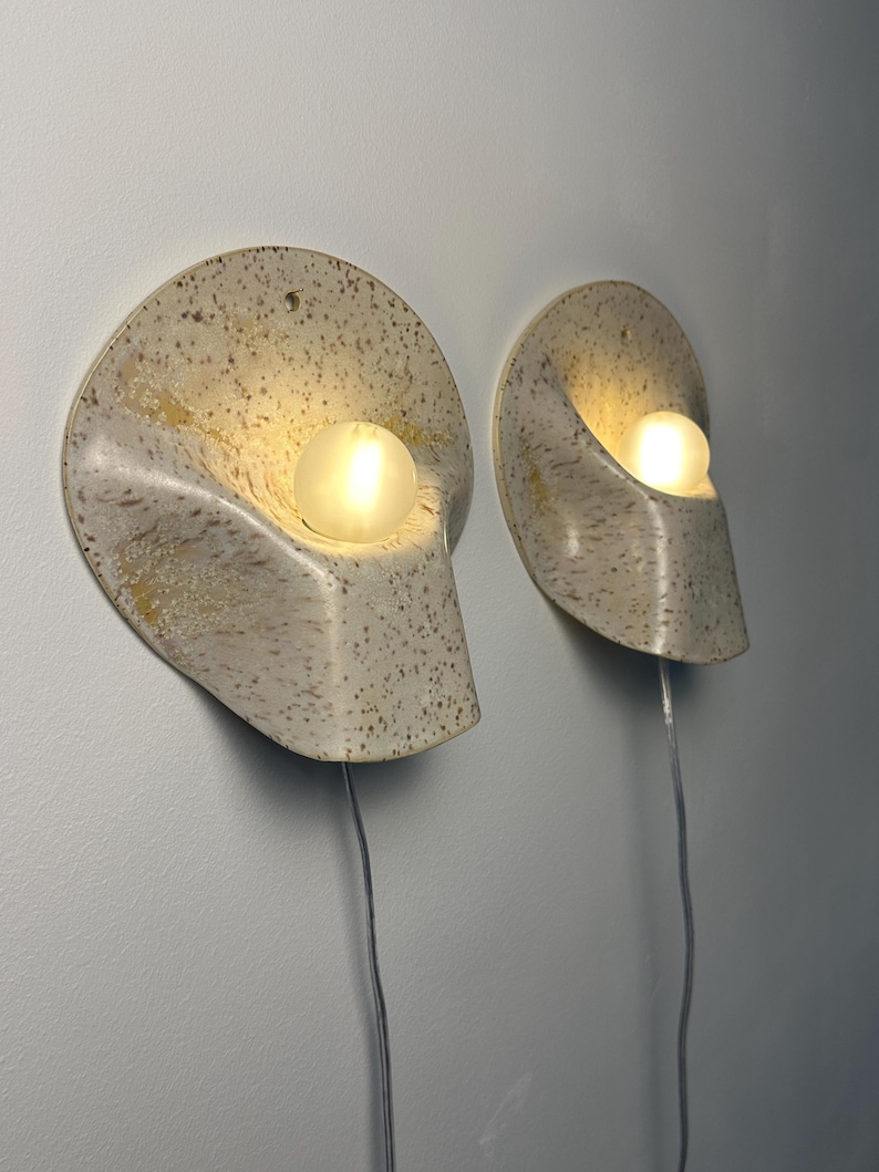 May include: Two wall-mounted sconces featuring a speckled cream and tan ceramic shell-like shade with a curved, organic form. Each fixture holds a single glowing spherical light bulb and includes a thin, clear electrical cord extending downwards.