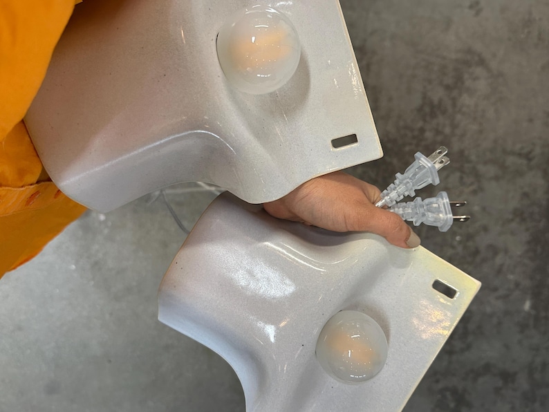 May include: Two white, curved, wall-mounted light fixtures with a glossy finish. Each fixture features a single, round, clear glass bulb and a rectangular slot near the edge. Two clear electrical cords with standard two-prong plugs are attached.
