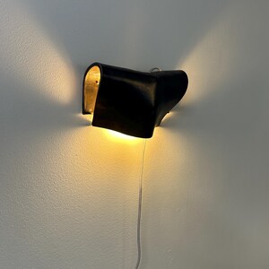 May include: A wall-mounted lamp featuring a curved, matte black exterior shell with a metallic gold interior finish. The fixture emits a warm, glowing light from both sides, casting beams upwards and downwards against the wall, with a thin power cord.