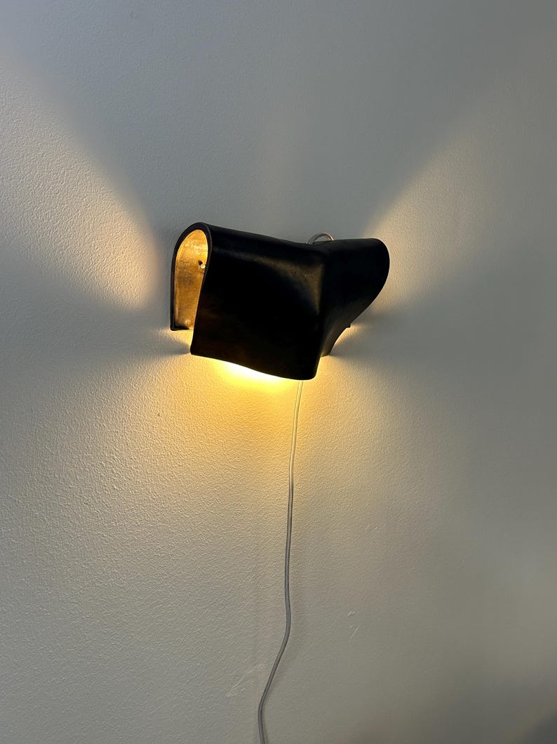 May include: A wall-mounted lamp featuring a curved, matte black exterior shell with a metallic gold interior finish. The fixture emits a warm, glowing light from both sides, casting beams upwards and downwards against the wall, with a thin power cord.