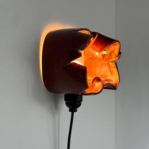 May include: A brown wall sconce with a unique, abstract design. The sconce is lit from within, casting a warm orange glow. A black cord extends from the base. The sconce is mounted on a white wall.