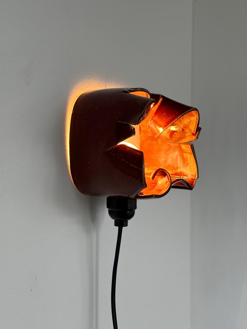 May include: A brown wall sconce with a unique, abstract design. The sconce is lit from within, casting a warm orange glow. A black cord extends from the base. The sconce is mounted on a white wall.