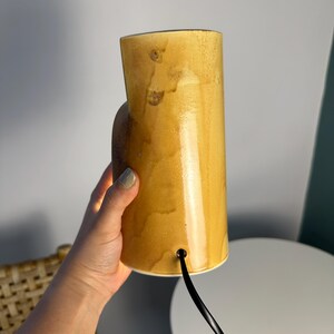Puede incluir: Una l&aacute;mpara cil&iacute;ndrica de cer&aacute;mica con un esmalte brillante de color amarillo mostaza moteado. La l&aacute;mpara presenta un cuerpo vertical liso con una peque&ntilde;a abertura circular cerca de la base por donde sale un cable el&eacute;ctrico negro, con acabado c&aacute;lido.