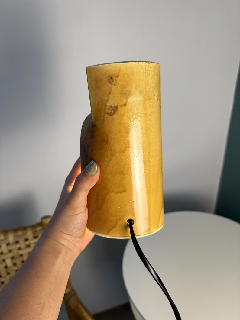 Puede incluir: Una l&aacute;mpara cil&iacute;ndrica de cer&aacute;mica con un esmalte brillante de color amarillo mostaza moteado. La l&aacute;mpara presenta un cuerpo vertical liso con una peque&ntilde;a abertura circular cerca de la base por donde sale un cable el&eacute;ctrico negro, con acabado c&aacute;lido.