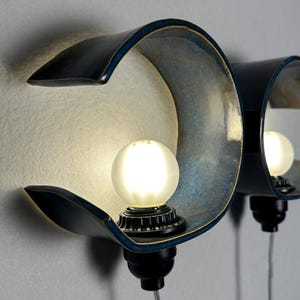 May include: Two wall-mounted sconces featuring curved, deep blue ceramic shades with a metallic interior finish. Each fixture holds a single frosted spherical light bulb centered within the arc, connected to a black base with a clear power cord.
