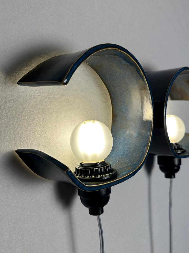 May include: Two wall-mounted sconces featuring curved, deep blue ceramic shades with a metallic interior finish. Each fixture holds a single frosted spherical light bulb centered within the arc, connected to a black base with a clear power cord.