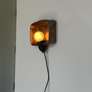 May include: A wall-mounted light fixture with a square, bronze-colored metal shade. The interior of the shade is gold, and it houses a round, glowing light bulb. A black cord extends downward from the fixture.