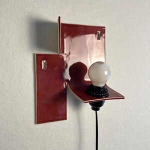 Pode incluir: Uma arandela de parede moderna com um design geom&eacute;trico. A arandela apresenta uma constru&ccedil;&atilde;o em azulejo cer&acirc;mico vermelho escuro e brilhante. Uma l&acirc;mpada branca assenta numa pequena prateleira, com base e cabo pretos. A lumin&aacute;ria &eacute; montada numa parede branca.