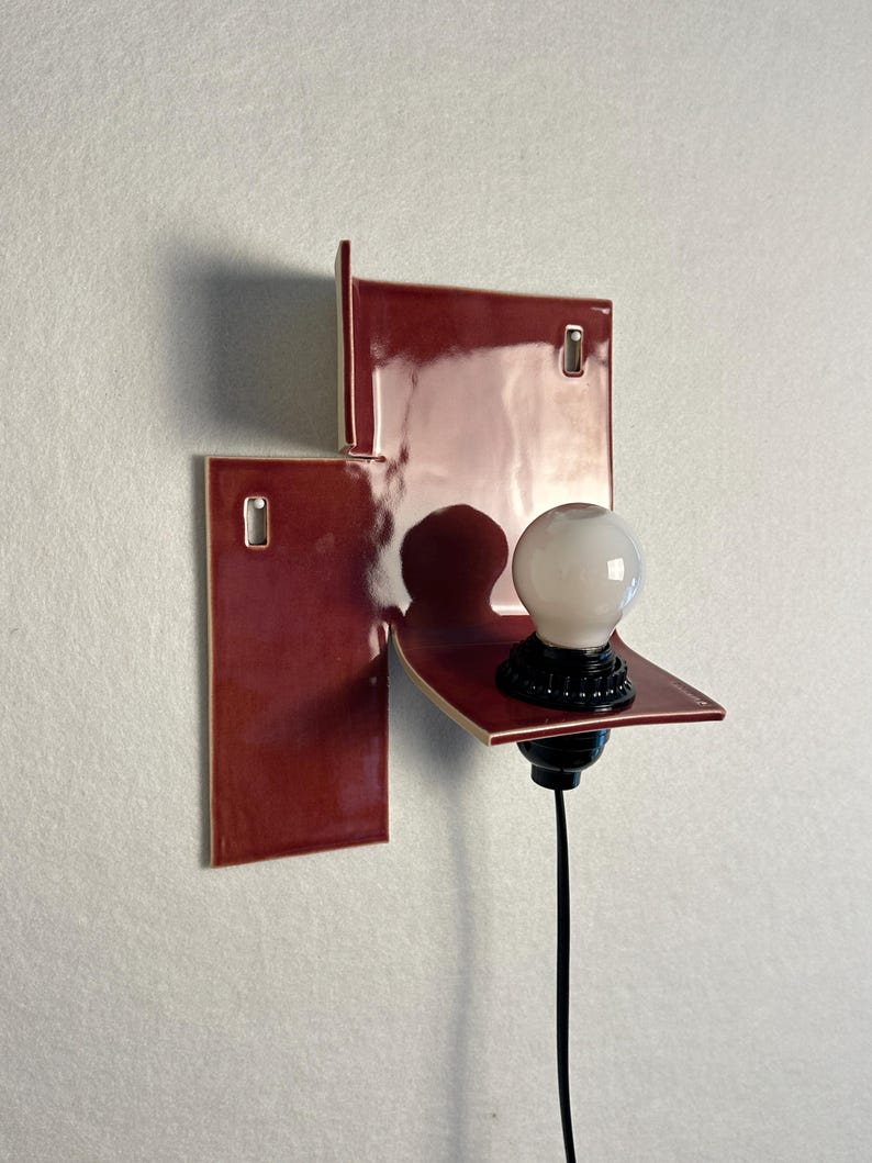 Pode incluir: Uma arandela de parede moderna com um design geom&eacute;trico. A arandela apresenta uma constru&ccedil;&atilde;o em azulejo cer&acirc;mico vermelho escuro e brilhante. Uma l&acirc;mpada branca assenta numa pequena prateleira, com base e cabo pretos. A lumin&aacute;ria &eacute; montada numa parede branca.