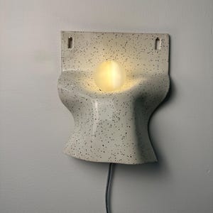 Może przedstawiać: Lampa ścienna z zakrzywionym, kremowym korpusem ceramicznym w czarne kropki. Oprawa posiada centralny, okrągły otw&oacute;r, w kt&oacute;rym znajduje się świecąca, kulista żar&oacute;wka. Z dolnej części urządzenia wychodzi czarny przew&oacute;d zasilający.