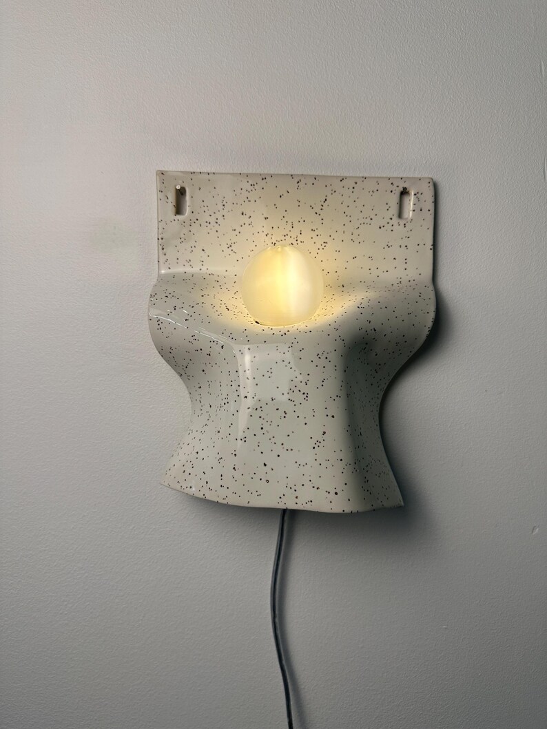 Może przedstawiać: Lampa ścienna z zakrzywionym, kremowym korpusem ceramicznym w czarne kropki. Oprawa posiada centralny, okrągły otw&oacute;r, w kt&oacute;rym znajduje się świecąca, kulista żar&oacute;wka. Z dolnej części urządzenia wychodzi czarny przew&oacute;d zasilający.