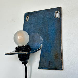May include: A modern wall sconce with a blue ceramic backplate and a black light fixture. The sconce features a frosted globe light bulb and a black electrical cord. The ceramic backplate has two rectangular cutouts for mounting.