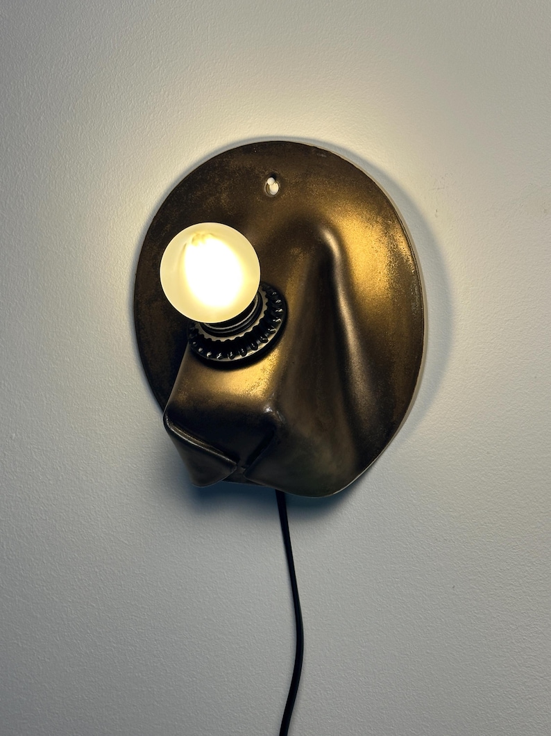 May include: A wall-mounted lamp featuring a circular, metallic bronze-toned backplate with a draped, fabric-like texture. A single round, glowing light bulb is secured in a black socket, and a thin black power cord extends downward from the base.