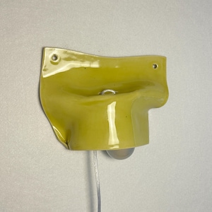 May include: A yellow wall-mounted light fixture with a bulb and a clear cord. The fixture has a unique, curved design with a small metal ring. The light is mounted on a neutral-colored wall.